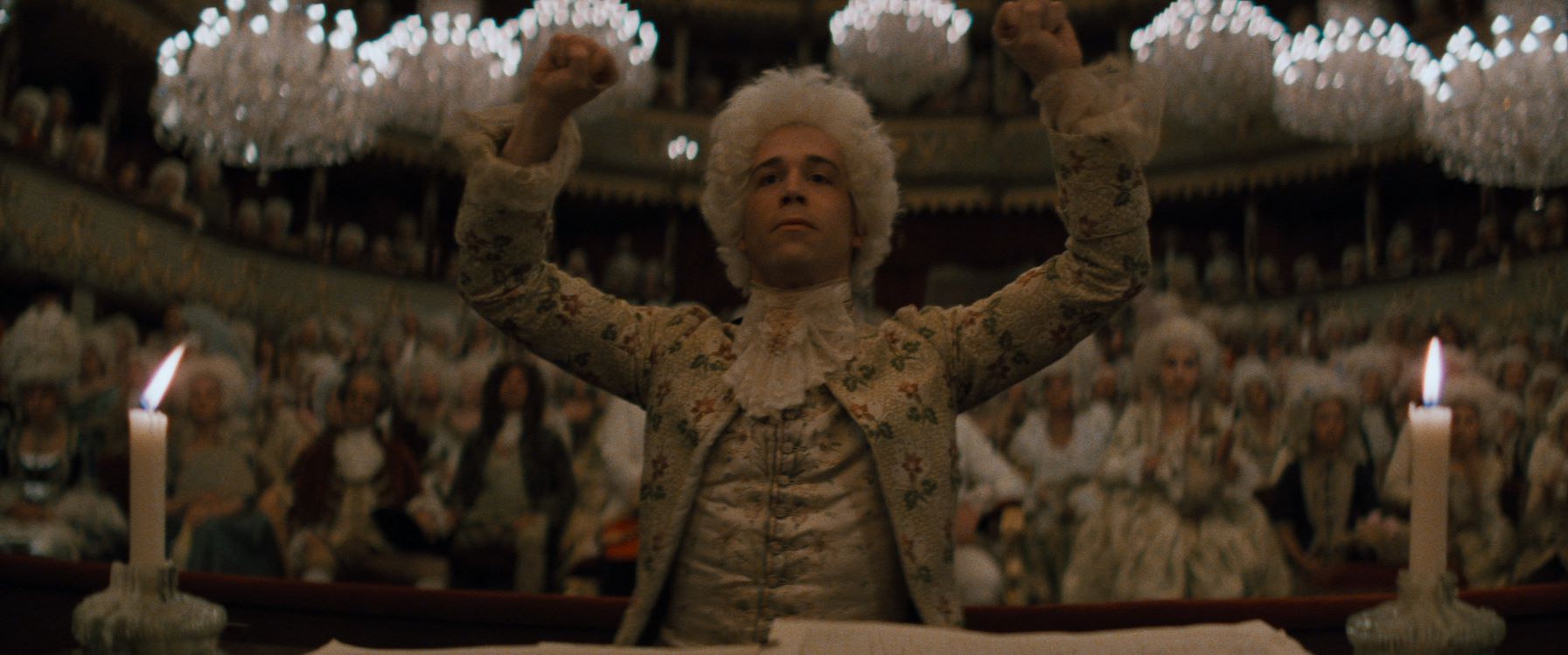 Amadeus Mozart is aan het dirigeren. Achter hem zien we een concertzaal. Iedereen heeft typisch witte hoge pruiken op.